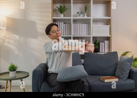 Ein junger Mann in einem grauen Hemd entspannt sich und dehnt seine Arme bequem, während er zu Hause auf einem gemütlichen blauen Sofa sitzt. Er genießt einen friedlichen Moment, lächelt Stockfoto
