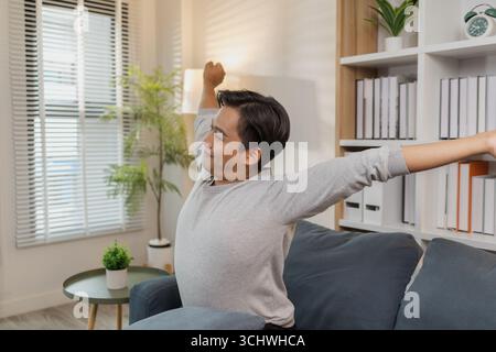Ein junger Mann in einem grauen Hemd entspannt sich und dehnt seine Arme bequem, während er zu Hause auf einem gemütlichen blauen Sofa sitzt. Er genießt einen friedlichen Moment, lächelt Stockfoto