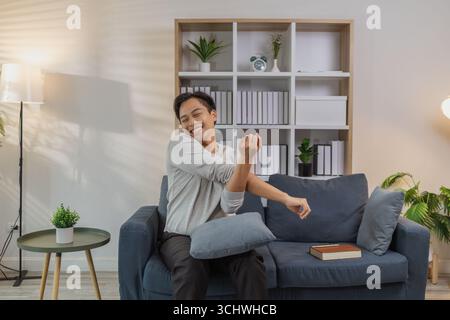 Ein junger Mann in einem grauen Hemd entspannt sich und dehnt seine Arme bequem, während er zu Hause auf einem gemütlichen blauen Sofa sitzt. Er genießt einen friedlichen Moment, lächelt Stockfoto