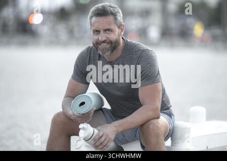 Sport-Fitness-Mann, der draußen auf einer Bank ruht. Reifes Fitnesssportmodell, das nach dem Training auf einer Bank sitzt. Senior Sports man macht danach eine Pause Stockfoto