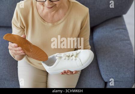 Ältere Frau sitzt zu Hause auf dem Sofa und setzt eine orthopädische Einlegesohle in einen Sneaker ein Stockfoto