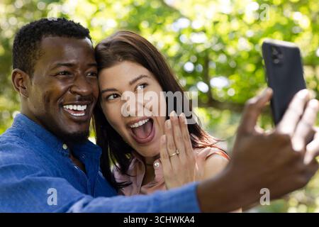Fröhliches, vielseitiges Paar, das im Freien Selfie macht und mit großem Lächeln die Verlobung feiert Stockfoto