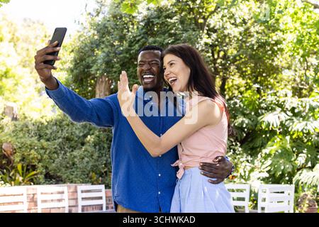 Glückliches, vielseitiges Paar, das Selfie im Freien macht und zusammen mit fröhlichen Ausdrücken feiert Stockfoto
