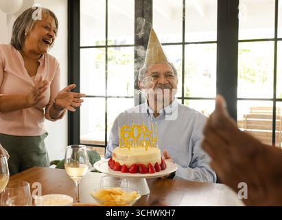 Wir feiern den 60. Geburtstag, Seniorenmann mit Kuchen und verschiedene Seniorenfreunde applaudieren zu Hause Stockfoto