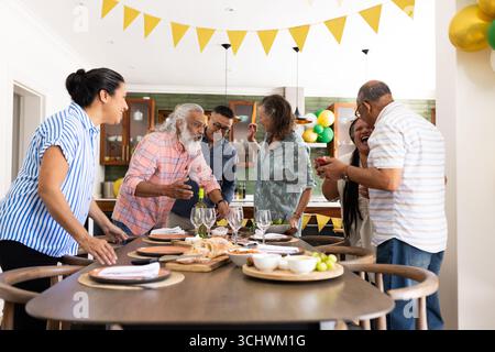 Verschiedene Seniorenfreunde lachen und plaudern am Esstisch während festlicher Feierlichkeiten zu Hause Stockfoto