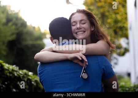 Junge, vielfältige Paare, die sich im Garten umarmen, eine Frau, die fröhlich lächelt, während sie die Schlüssel hält Stockfoto