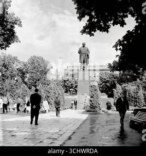 Ein Archivfoto in Schwarzweiß, aufgenommen um 1965 in einer großen Stadt auf der Krim (vermutlich Simferopol oder Jalta). Eine monumentale Statue von V.I. Lenin in einem zentralen Stadtpark nach einem Regenschauer. Sowjetische Bürger schlendern auf den nassen Pfaden. Das Foto spiegelt das tägliche Leben und die ideologischen Symbole einer sowjetischen Stadt in den 1960er Jahren wider, lange vor dem Prozess der Dekommunisierung in der Ukraine Stockfoto