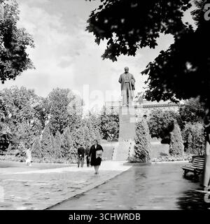 Ein Archivfoto in Schwarzweiß, aufgenommen um 1965 in einer großen Stadt auf der Krim (vermutlich Simferopol oder Jalta). Eine monumentale Statue von V.I. Lenin in einem zentralen Stadtpark nach einem Regenschauer. Sowjetische Bürger schlendern auf den nassen Pfaden. Das Foto spiegelt das tägliche Leben und die ideologischen Symbole einer sowjetischen Stadt in den 1960er Jahren wider, lange vor dem Prozess der Dekommunisierung in der Ukraine Stockfoto