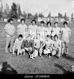 Eine Schwarzweißaufnahme aus dem Archiv, aufgenommen in Sviatohirsk in den 1970er Jahren Eine lokale Amateurmannschaft posiert für ein Gruppenfoto, nachdem sie ein Turnier gewonnen hat. Die Spieler und ihr Trainer stehen auf einem ländlichen Fußballfeld und halten ihre Auszeichnungen – Zertifikate und eine Trophäe. Das Foto vermittelt einen Geist der Kameradschaft und die Freude am Sieg, was die Kultur des Amateursports in der UdSSR widerspiegelt Stockfoto