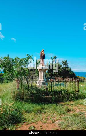 Statue des Heiligen Antonius von Padua, der ein Kind auf einem Steinsockel hält, umgeben von einem rostigen Eisenzaun auf einem grasbewachsenen Feld auf der Ile Saint-Honorat, Cannes Stockfoto
