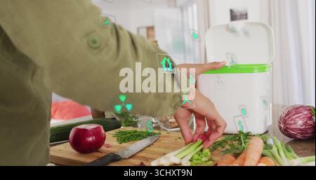 Hauskoch in grüner Jacke, der Gemüsereste in einen grünen Kompostbehälter auf der Küchentheke hebt Stockfoto
