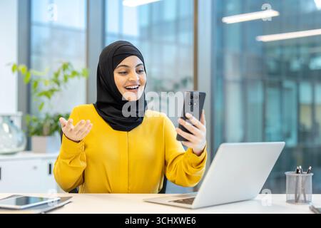 Die junge, fröhliche muslimische Geschäftsfrau im Hijab, die Videoanrufe per Smartphone hat, während sie im modernen Büro am Schreibtisch sitzt, macht Gesten und lächelt Stockfoto