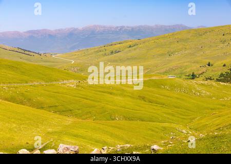 Gewundene Straße über das grüne Creueta-Plateau in den Pyrenäen mit weichen Hügeln, felsigem Gelände und weit entfernten Berggipfeln unter klarem blauen Himmel. Stockfoto
