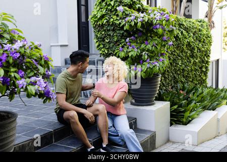 Junges, vielfältiges Paar, das auf Stufen sitzt, lacht und die gemeinsame Zeit auf der Terrasse genießt Stockfoto