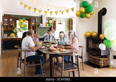 Feiern Sie mit Ballons, verschiedene Seniorenfreunde, die zusammen zu Mittag essen und sich zu Hause unterhalten Stockfoto