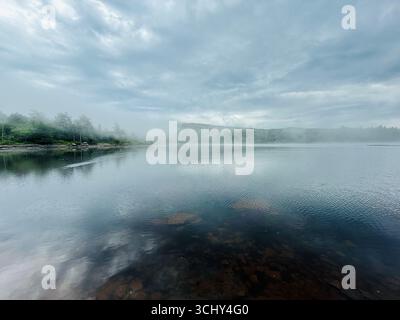 Dichter, dicker, grauer Sonnenaufgangsnebel und Morgennebel rollt über dem Wasser am North South Lake in Haines Falls, New York. Bäume, Sonne, hohe Berge hinter Wolken Stockfoto