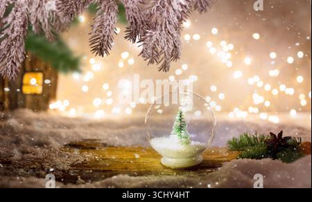 Dekorativer Tannenbaum mit Kunstschnee in einer Glas-Weihnachtsbaumkugel, Holzhaus und Lichter im Hintergrund Stockfoto