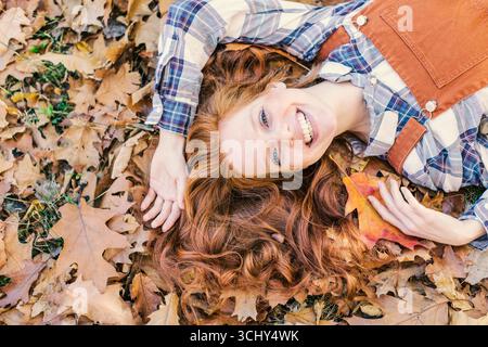 Rothaarige Frau, die glücklich im Herbstlaub liegt und in die Kamera blickt Stockfoto