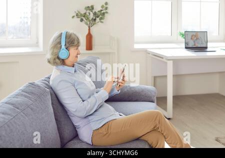 Ältere Frau im Kopfhörer, die Musik über das Smartphone hört und zu Hause auf dem Sofa sitzt. Stockfoto