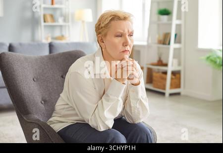 Eine nachdenkliche Seniorin sitzt zu Hause im Sessel, legt ihr Kinn auf die Hände und reflektiert tief. Stockfoto