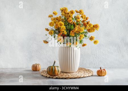 Herbstgelbe und orangene Blüten in weißer Vase und Kürbisse auf heller Oberfläche. Minimalistische moderne Komposition. Inneneinrichtung und Dekoration der Saison. Stockfoto
