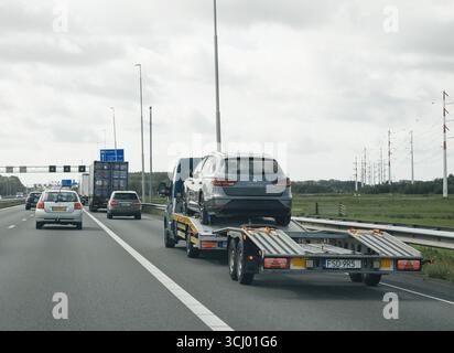 Haag, Niederlande - 26. August 2024: Ein Auto, das auf einem Anhänger beladen ist, wird auf einer niederländischen Autobahn transportiert, umgeben von Verkehr und bewölktem Wetter. Stockfoto