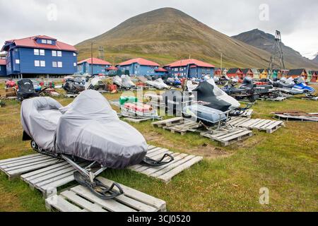 Schneemobile abgedeckt auf Paletten in Longyearbyen Svalbard Norwegen | LONGYEARBYEN, Svalbard — Yamaha und andere Schneemobile liegen abgedeckt auf Holzpaletten vor den farbcodierten Gebäuden von Longyearbyen, einem System, das historisch verwendet wurde, um die Funktion einer Struktur in der Kohlengemeinde zu kennzeichnen, die von Store Norske Spitsbergen Kulkompani betrieben wird. Ein steiler, kahler Berg erhebt sich hinter der Siedlung. Schneemobile bleiben ein primäres Beförderungsmittel über Land im Archipel während des langen, dunklen arktischen Winters. Stockfoto