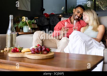 Vielfältiges Paar, das sich auf dem Sofa mit Wein entspannt und einen gemütlichen Nachmittag zu Hause genießt Stockfoto