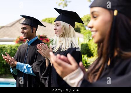 Verschiedene Absolventen in Mützen und Kleidern klatschen im Garten, um akademische Leistungen zu feiern Stockfoto