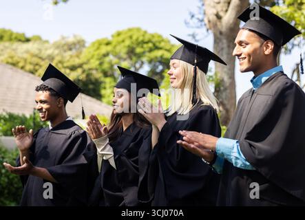Verschiedene Absolventen in Mützen und Kleidern klatschen im Garten und feiern ihre Leistung Stockfoto
