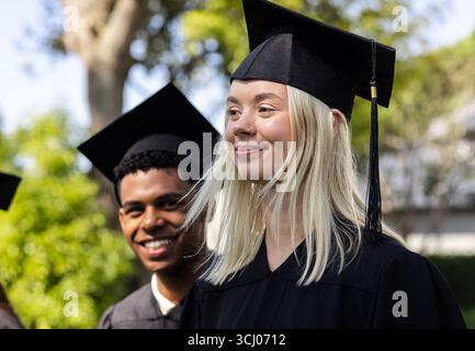 Absolventen verschiedener Studenten, die Kappen und Kleider tragen, lächelnd im Garten, feiern Erfolge Stockfoto