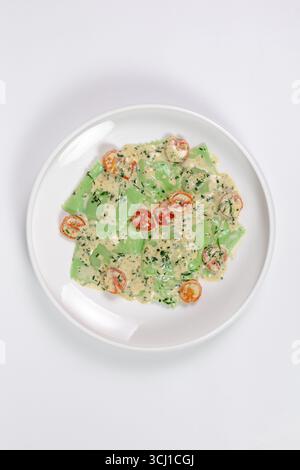 Frische grüne Ravioli aus mit Kräutern angereicherten Nudeln, serviert mit cremiger Käsesauce, halbierten Kirschtomaten und gehackten Kräutern auf einem weißen Teller. Minimal Stockfoto