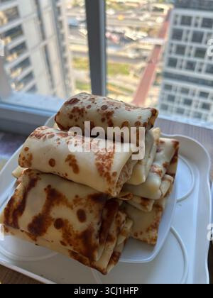 Das Bild zeigt einen Stapel goldbrauner, gefalteter Pfannkuchen, die auf einer weißen Plastikplatte auf einer Fensterbank angeordnet sind. Im Hintergrund sehen Sie die Stockfoto