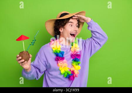 Junger Mann, der ein tropisches Getränk hält, trägt bunten lei und Strohhut mit aufgeregtem Ausdruck auf grünem Hintergrund Stockfoto