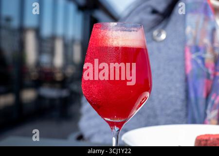 Erfrischender roter Cocktail in elegantem Glas mit Blasen und Kondenswasser, der auf einem Tisch mit einer verschwommenen Essensszene im Freien platziert wird, sorgt für eine lebendige Atmosphäre Stockfoto