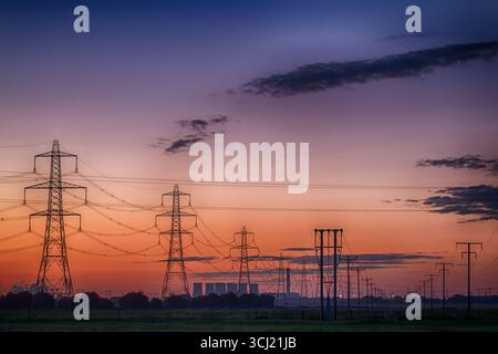 Reihen von Strommasten und Stromleitungen, die sich bei Sonnenaufgang in die Ferne erstrecken, mit einem Kraftwerk und Kühltürmen am Horizont Stockfoto