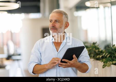 Lächelnder Geschäftsmann mittleren Alters im Anzug, der vor dem Büro steht und ein digitales Tablet hält. Stockfoto