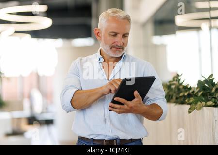 Lächelnder Geschäftsmann mittleren Alters im Anzug, der vor dem Büro steht und ein digitales Tablet hält. Stockfoto