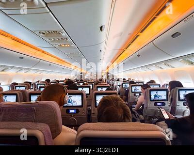 emirates Boeing 777 Flugzeugkabine gefüllt mit Bildschirmen auf der Rückseite der Sitze während eines Tagesflugs mit orangefarbener Umgebungsbeleuchtung Stockfoto