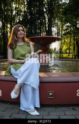 Eine junge Frau sitzt an einem Brunnen in einem Park, hält ein offenes Buch und blickt nachdenklich in die Ferne Stockfoto