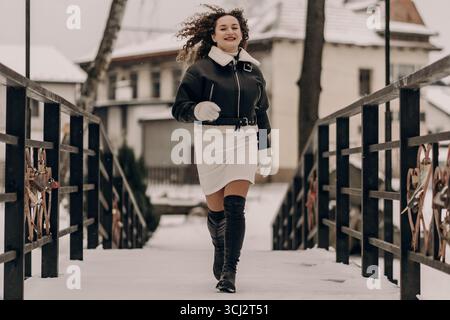 Eine Frau mit lockigen Haaren steht in einem verschneiten Stadtgebiet und trägt eine warme Jacke. Dunkle Stadtstrukturen umgeben sie, die den Kontrast von Kälte hervorheben Stockfoto
