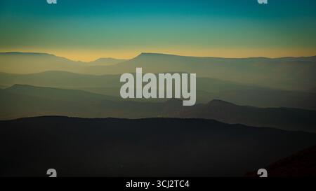 Neblige marokkanische Berglandschaft mit mehrschichtigen Bergrücken, die in sanftem Licht leuchten, und atmosphärischem Dunst, der sich über den Horizont erstreckt Stockfoto