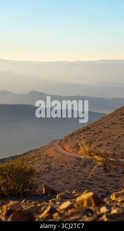 Felsiger marokkanischer Berghang mit Wüstenpflanzen und gewundenem Schotterweg mit Blick auf die neebedeckten Bergrücken unter der warmen Abendsonne Stockfoto