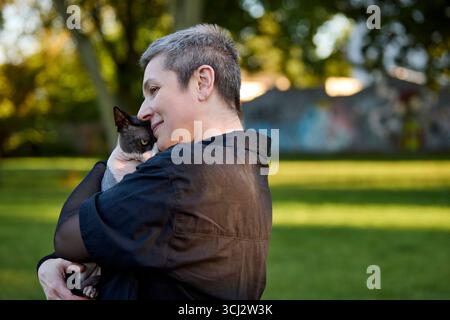 Eine Frau mit abgeschnittenem grauem Haar umarmt ihre kanadische Sphynx-Katze in einem sonnigen Park. Das Bild vermittelt Zärtlichkeit, Komfort und die starke emotionale c Stockfoto