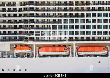 Nahaufnahme des Außenbereichs eines Kreuzfahrtschiffes mit mehreren Deck und orangefarbenen Rettungsbooten und Kabinen mit Balkonen und reflektierenden Fenstern. Stockfoto
