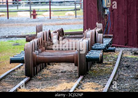 Eine Sammlung rostiger Zugräder befindet sich auf Eisenbahngleisen neben einer Scheune. Die Umgebung hat eine ländliche Atmosphäre mit Kies und grünem Gras im Hintergrund. Stockfoto