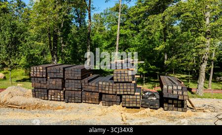 Ein großer Stapel Holz ist ordentlich auf einer Baustelle angeordnet, die von grünen Bäumen umgeben ist. Das helle Sonnenlicht hebt die robusten Holzbalken hervor und zeigt die Vorbereitung auf Bauprojekte an. Stockfoto