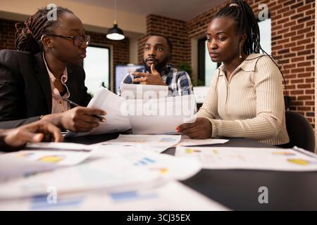 afroamerikanische Unternehmer mit Dokumenten, in denen Besprechungen und Beratungen rund um Besprechungstische in Backsteinmauerbüros diskutiert werden. Das Startup-Team plant gemeinsam Geschäftsprojekte und arbeitet an einer Vereinbarung. Stockfoto