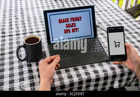 Eine Person sitzt mit einer Tasse Kaffee an einem Tisch und nutzt einen Laptop, um die Black Friday-Verkäufe zu durchsuchen. Sie halten eine Kreditkarte in der einen Hand und ein Smartphone mit einem Einkaufswagen in der anderen Hand. Stockfoto