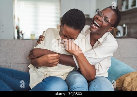 Zwei junge schwarze Frauen, die zu Hause auf der Couch lachen und umarmen Stockfoto